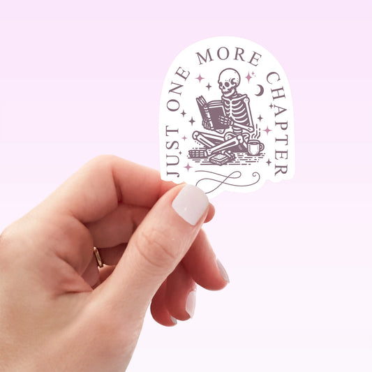 Hand holding a 'Just One More Chapter' sticker with a skeleton reading a book design on a light purple background.