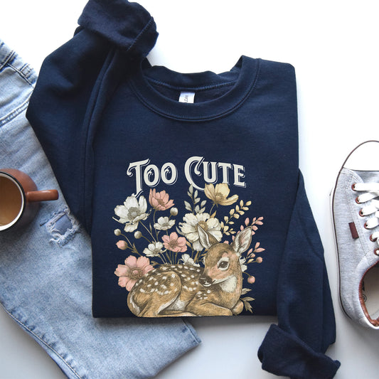 Navy blue sweatshirt with a deer and floral design labeled 'Too Cute' on a white surface with denim jeans and a tennis shoe.
