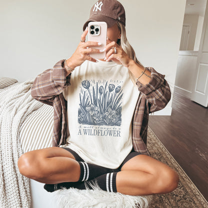Person wearing a white t-shirt with a wildflower graphic design, plaid shirt, and NY cap sitting on a couch.