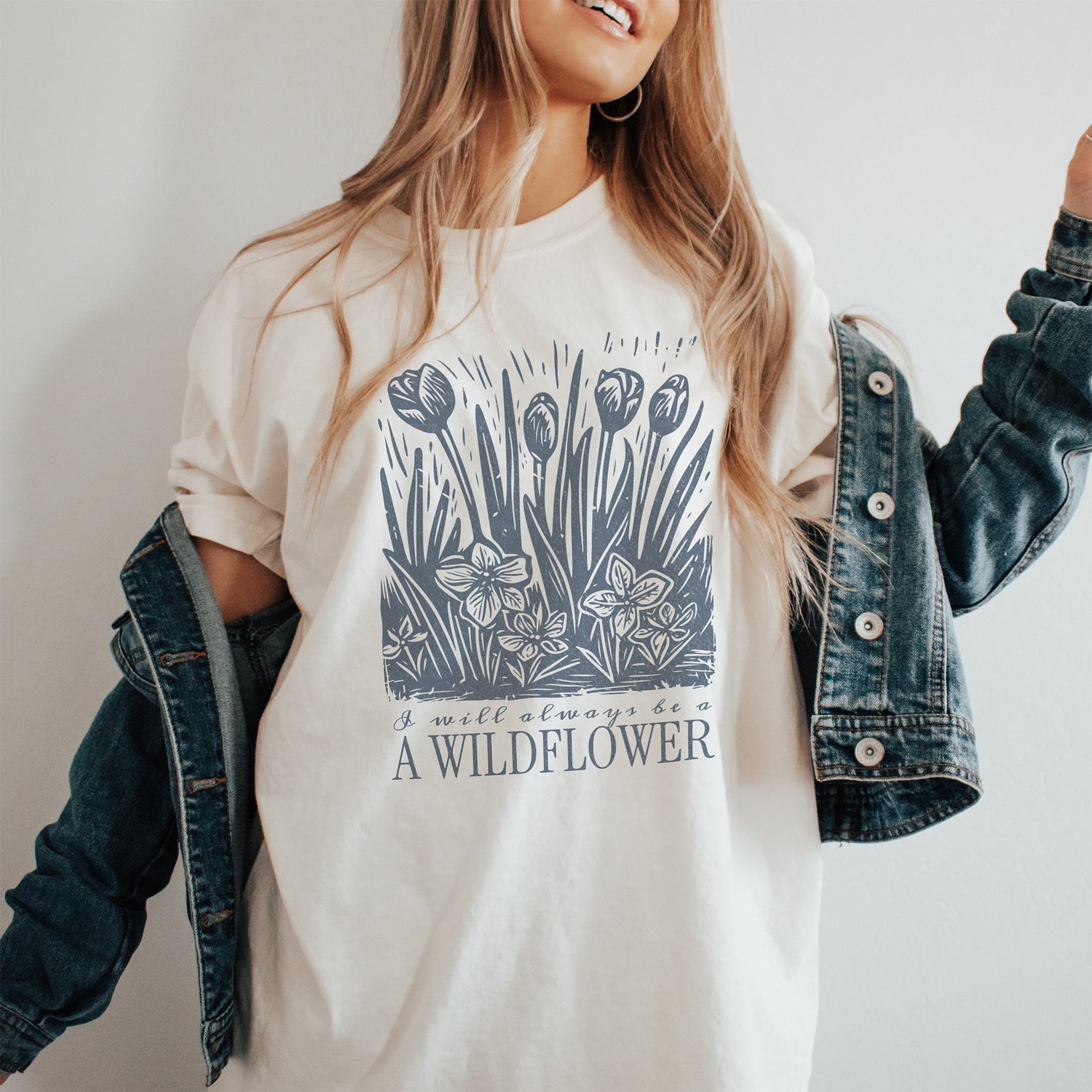 Person wearing a white t-shirt with a floral design and text that says I will always be a wildflower, paired with a denim jacket.