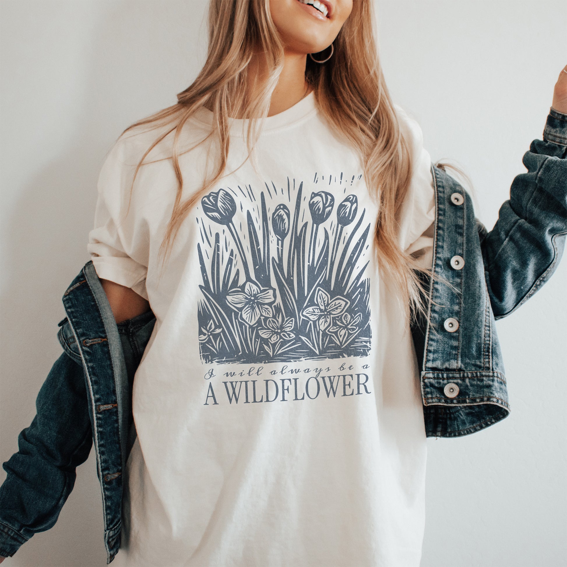 Person wearing a white t-shirt with a floral design and text that says I will always be a wildflower, paired with a denim jacket.