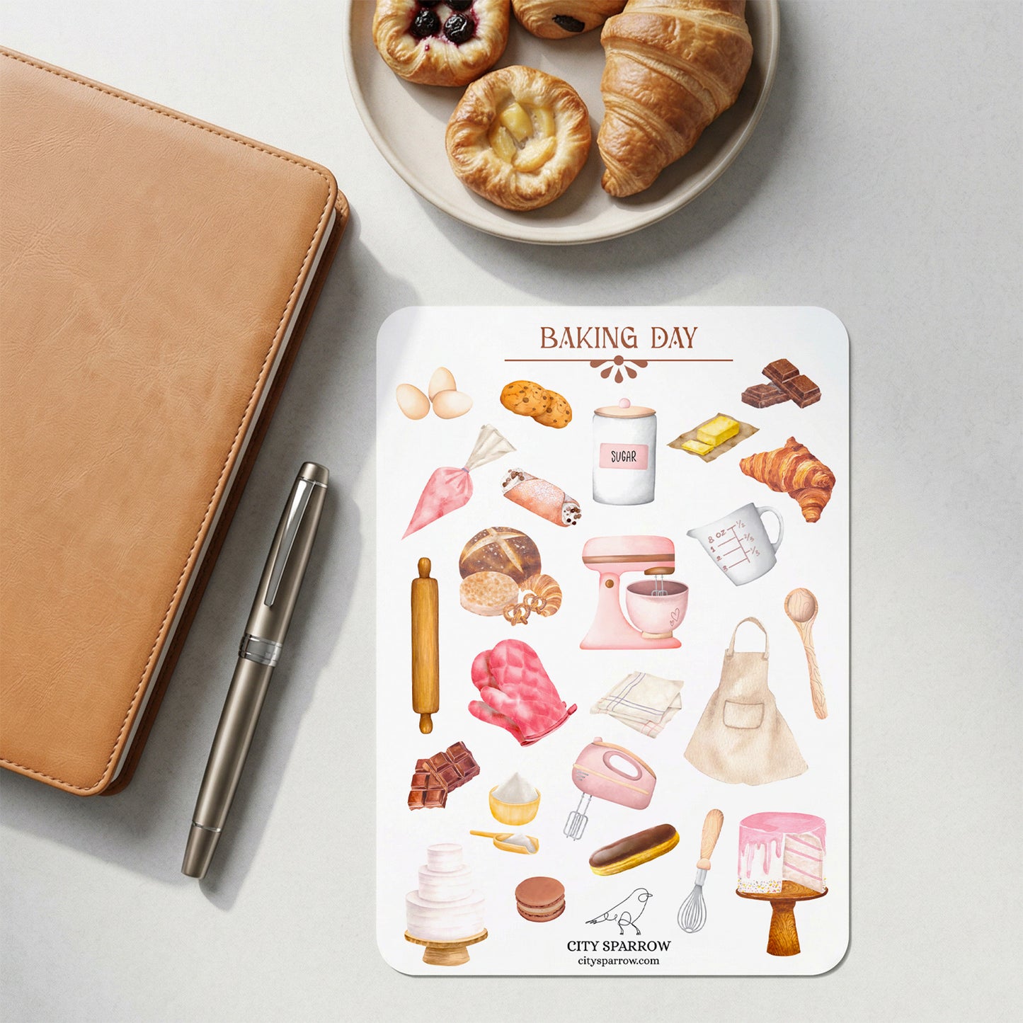 Sticker sheet with baking-themed illustrations on a white surface with a pen and plate of pastries.