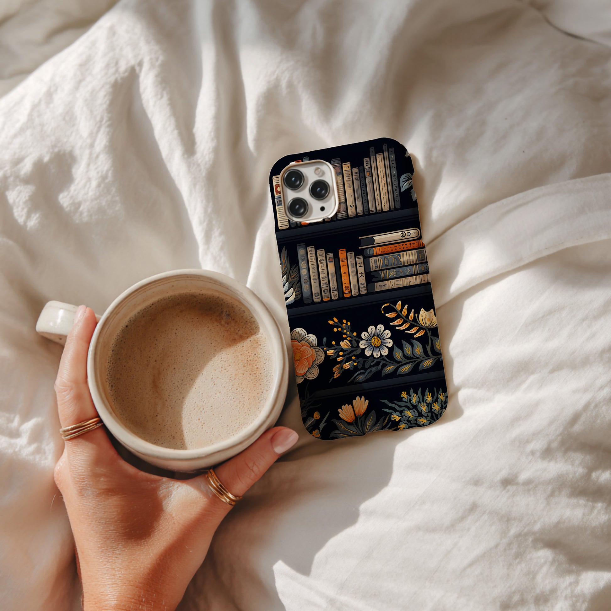 Person holding a mug with a phone case featuring book and floral design on a soft fabric background