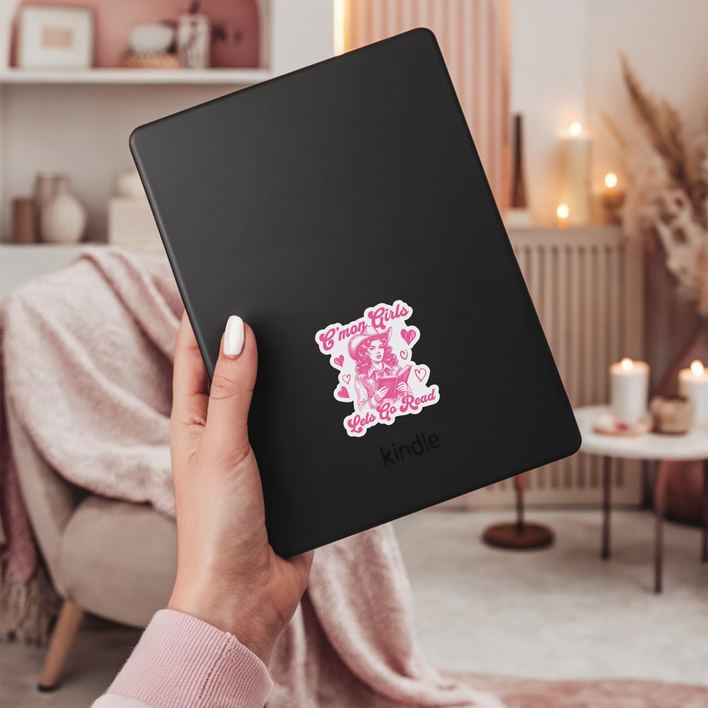Person holding a black Kindle with a sticker with a pink and white design a cowgirl and the text C'mon girls, let's go read on a blurred indoor background
