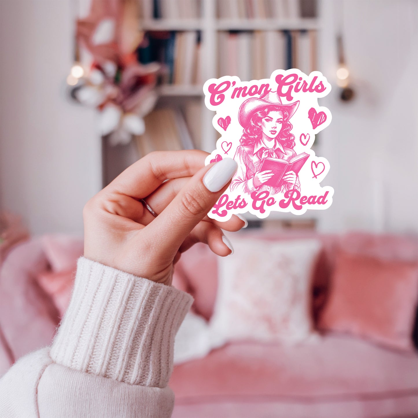 Hand holding a pink sticker with text that says C'mon girls, let's go read and a cowgirl  against a blurred indoor background