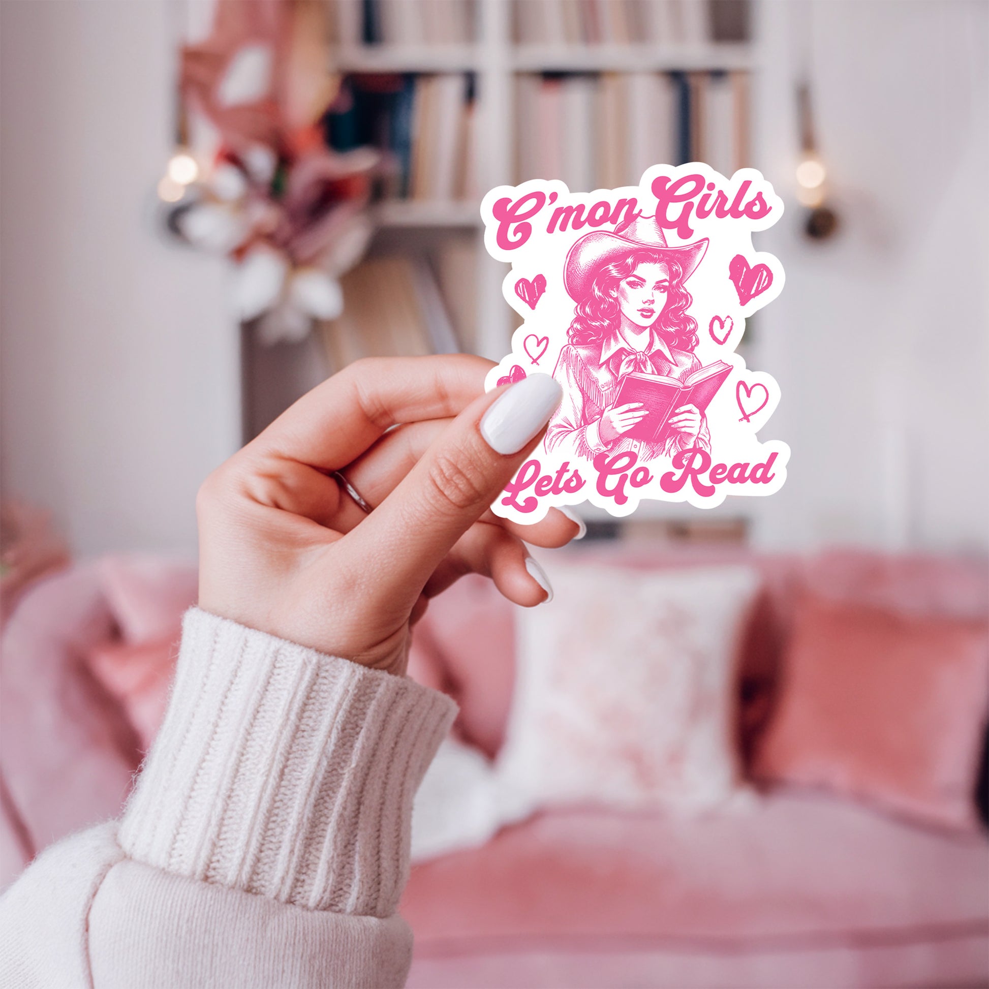 Hand holding a pink sticker with text that says C'mon girls, let's go read and a cowgirl  against a blurred indoor background