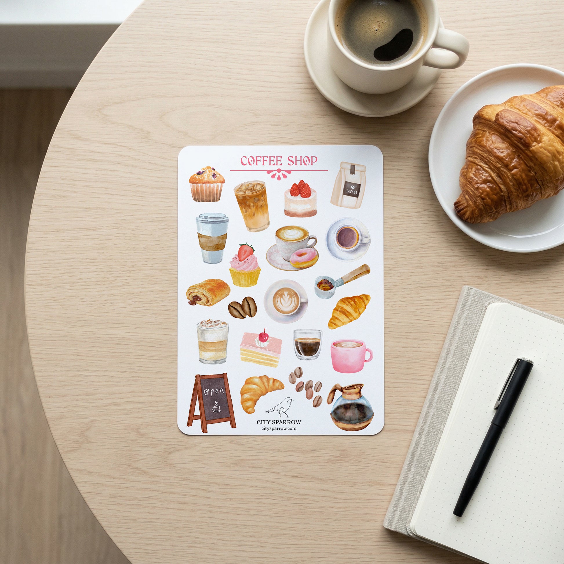Sticker sheet with coffee shop-themed illustrations on a table with a cup of coffee and croissant.