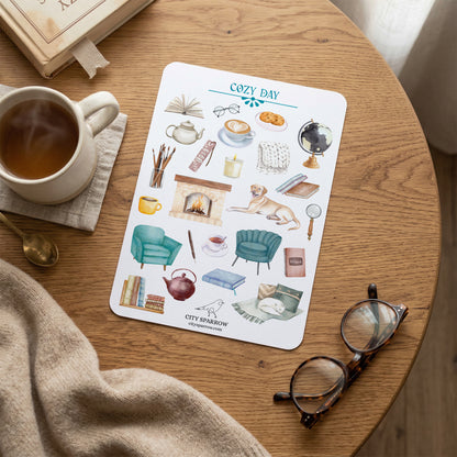 Sticker sheet titled 'Cozy Day' on a wooden table with a cup of tea, glasses, and a blanket.