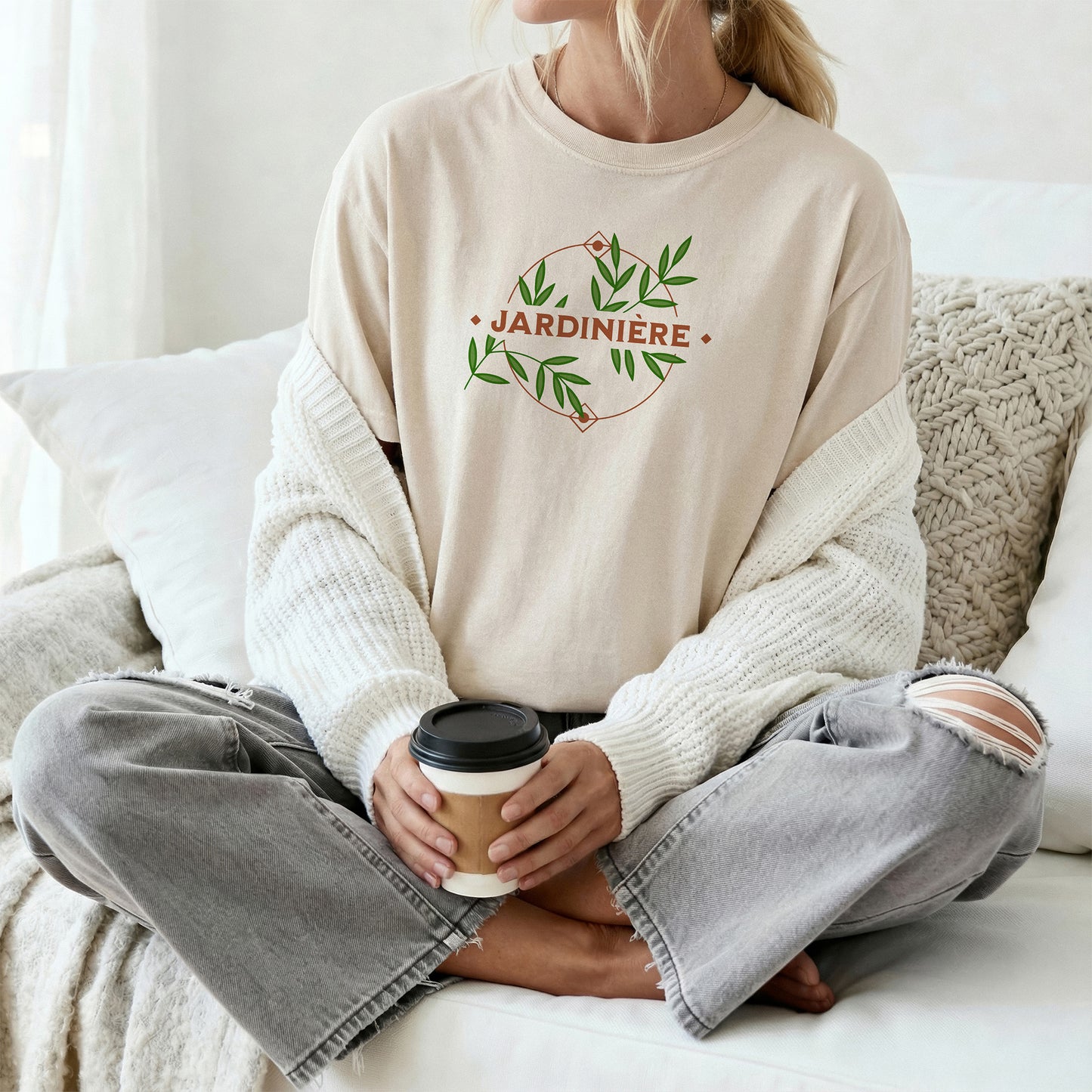 Person wearing a beige tee shirt with 'Jardinière' print, holding a coffee cup on a couch.
