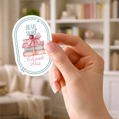 Hand holding a sticker with book-themed design and text that says Read More Tolerate Less  with a stack of books tied with a bow