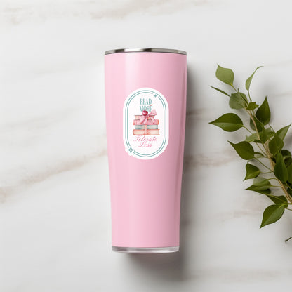 Pink tumbler with a Hand holding a sticker with book-themed design and text that says Read More Tolerate Less  with a stack of books tied with a bow
