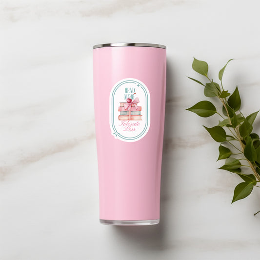 Pink tumbler with a Hand holding a sticker with book-themed design and text that says Read More Tolerate Less  with a stack of books tied with a bow