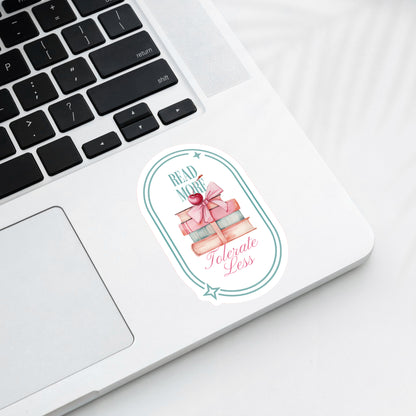 A sticker with book-themed design and text that says Read More Tolerate Less  with a sick of books tied with a bow pictured on a laptop