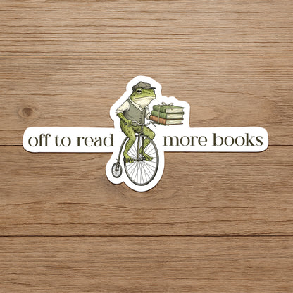 Frog on a bicycle with books, text 'off to read more books' on a wooden background