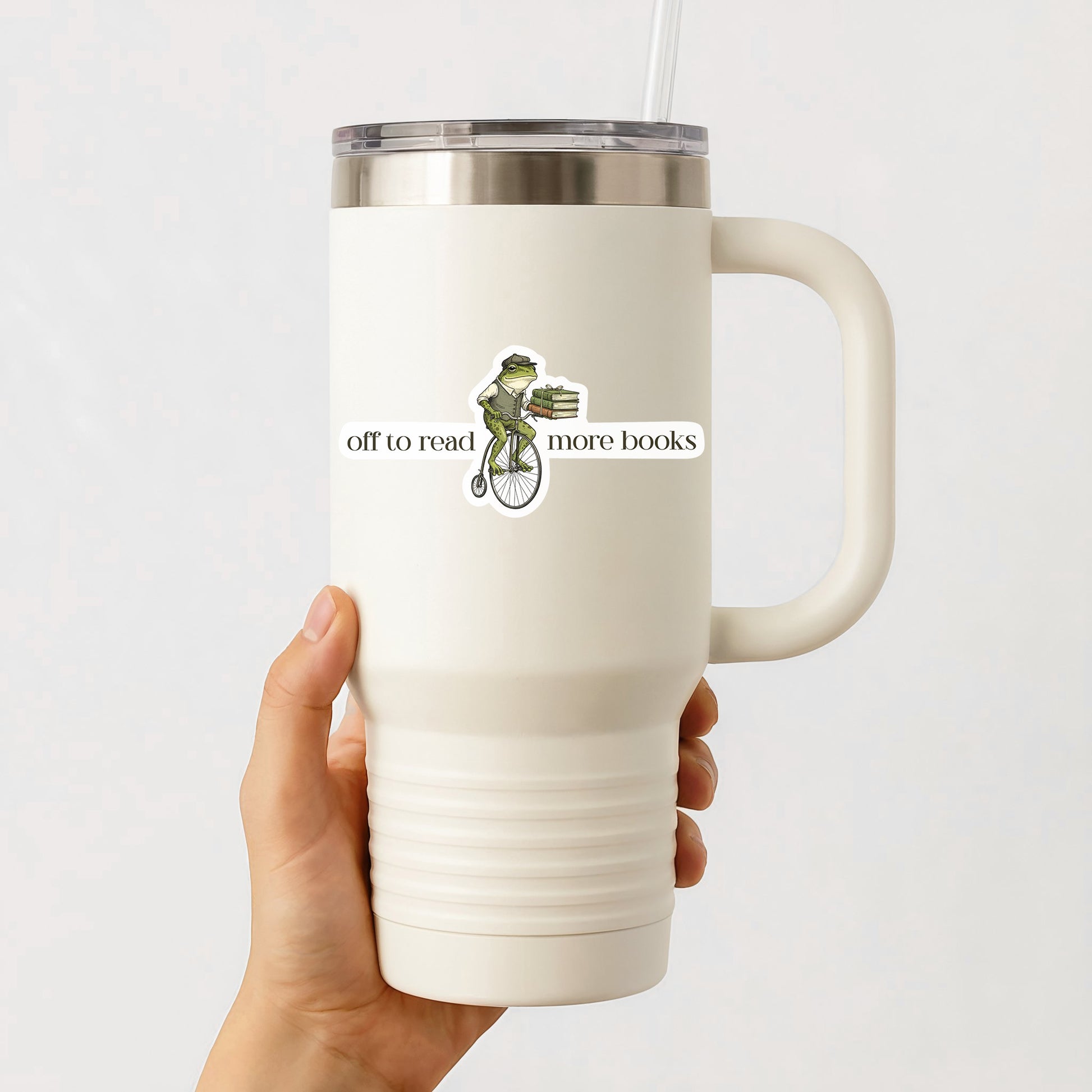 White travel mug with a handle held by a hand, featuring a frog sticker with text that says off to read more books on a white background.