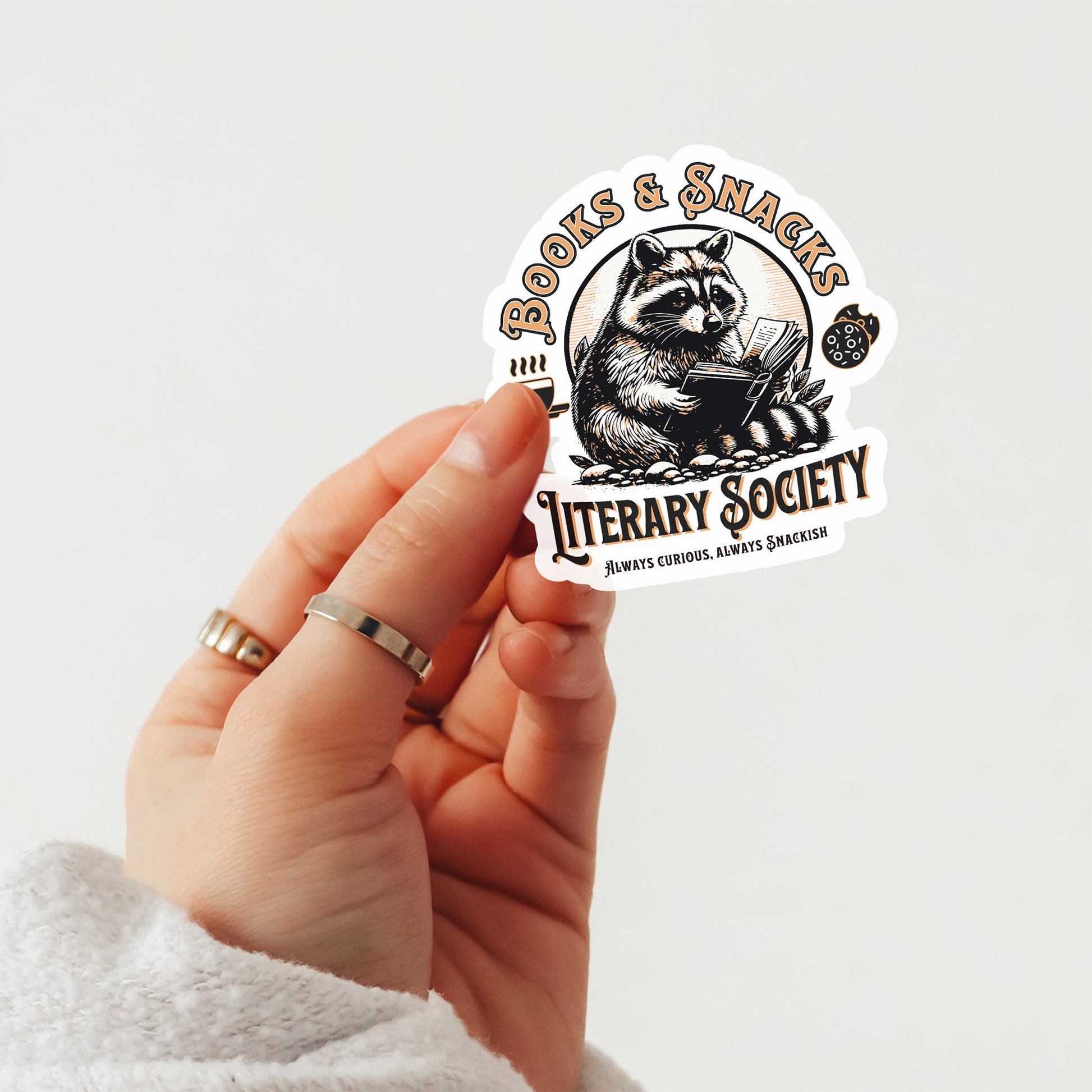 Hand holding a sticker with a raccoon design and text saying books and snacks literary society on a light gray background