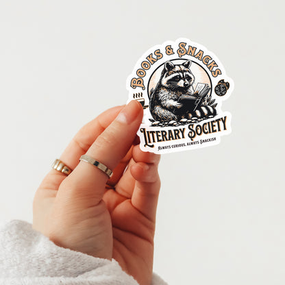 Hand holding a sticker with a raccoon design and text saying books and snacks literary society on a light gray background