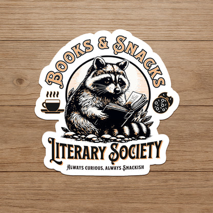 Sticker with a raccoon reading a book and holding a snack, labeled 'Books & Snacks Literary Society' on a wooden background.