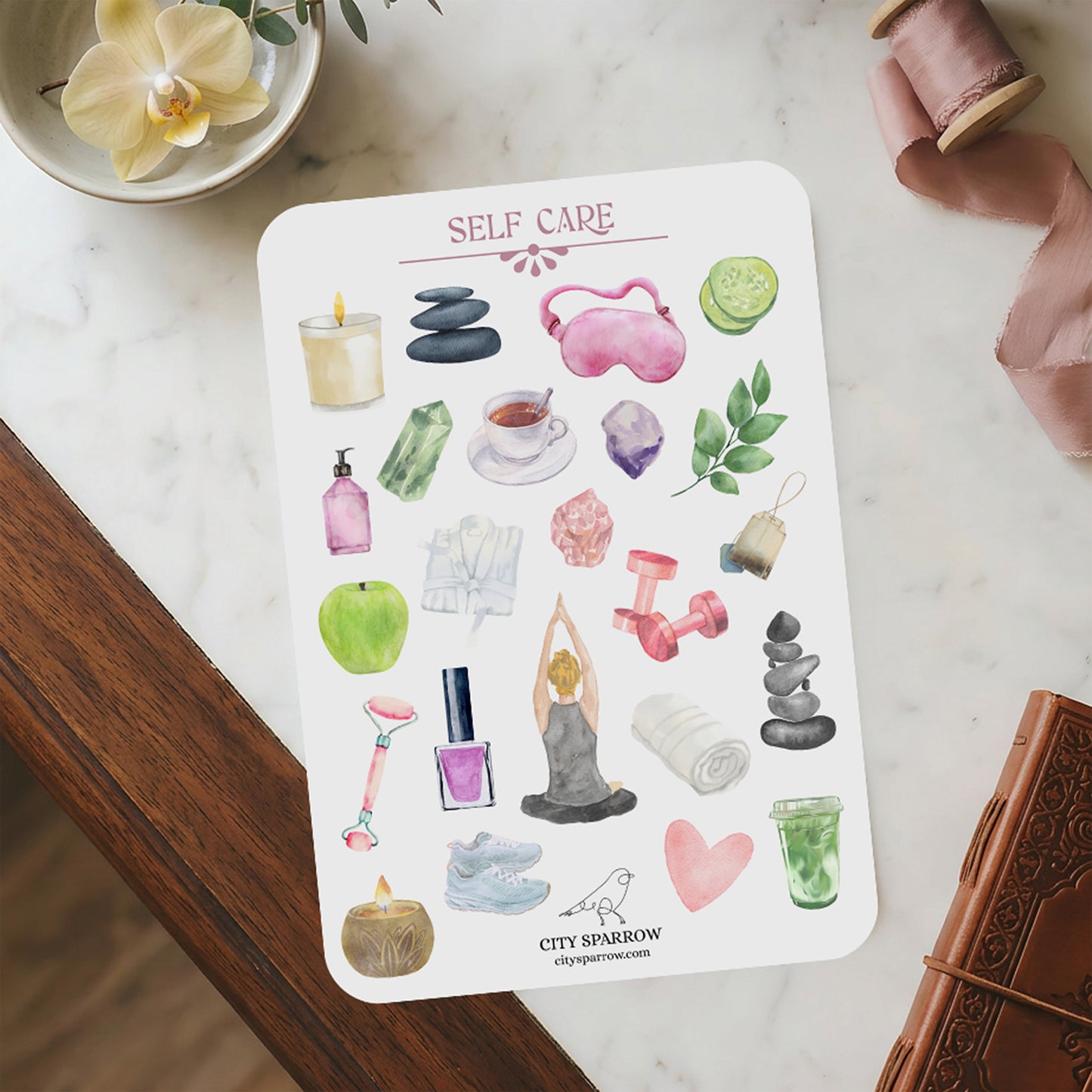 Sheet of self-care stickers on a marble surface with a small plant and book.