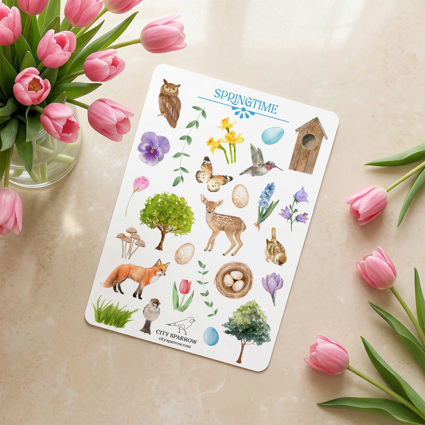 Sheet of spring-themed stickers with flowers, animals, and nature elements on a beige surface with pink tulips.