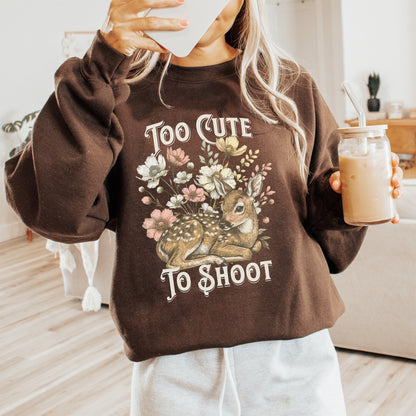 Person holding a drink and wearing a brown sweatshirt with a deer graphic and text saying too cute to shoot.