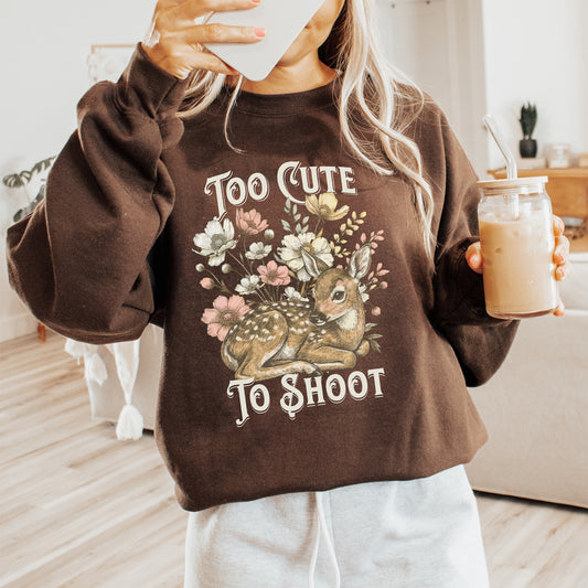 Person holding a drink and wearing a brown sweatshirt with a deer graphic and text saying too cute to shoot.