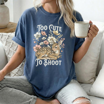 Person wearing a blue short sleeved t-shirt with a deer and floral design with text reading too cute to shoot while sitting on a couch holding a mug.