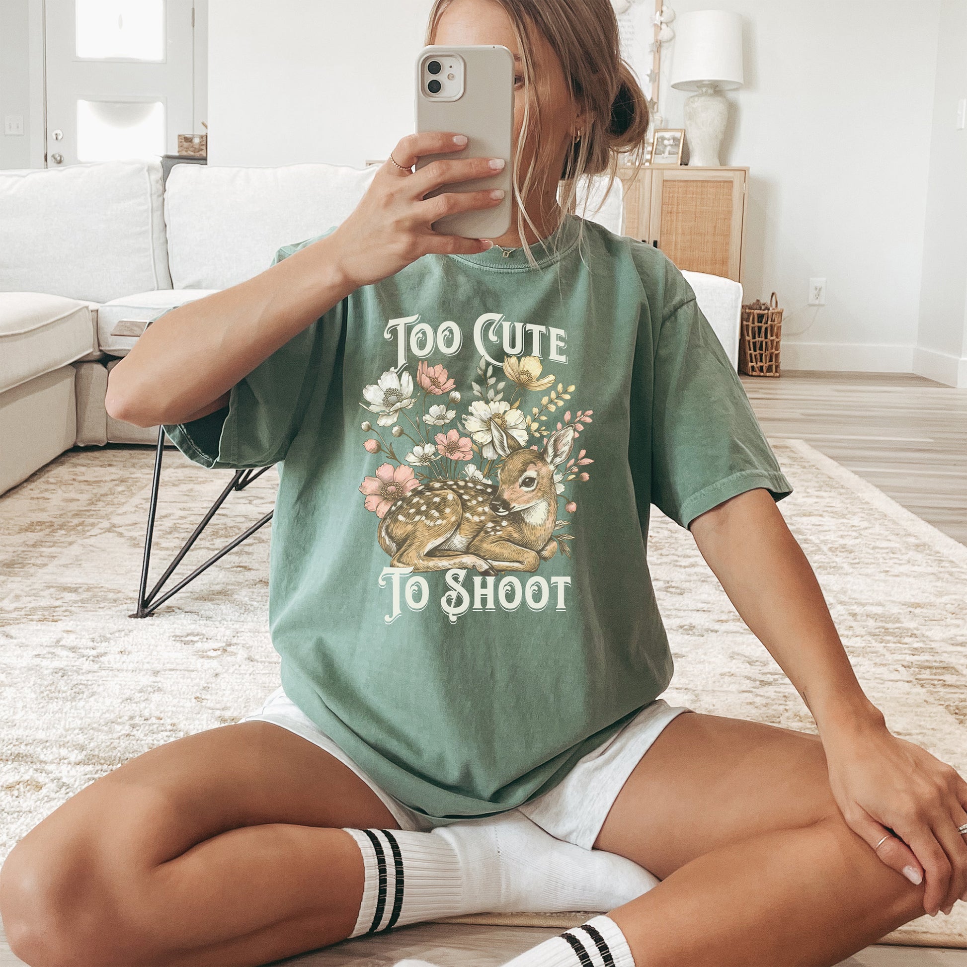 Woman wearing a green t-shirt with a deer graphic and text reading too cute to shoot while sitting on the floor of a light colored living room.