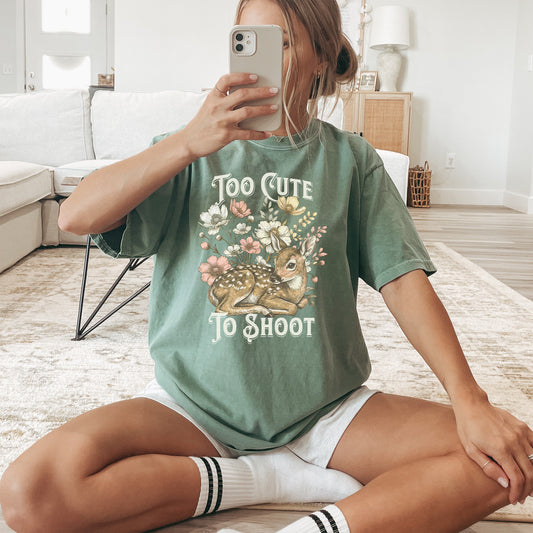 Woman wearing a green t-shirt with a deer graphic and text reading too cute to shoot while sitting on the floor of a light colored living room.