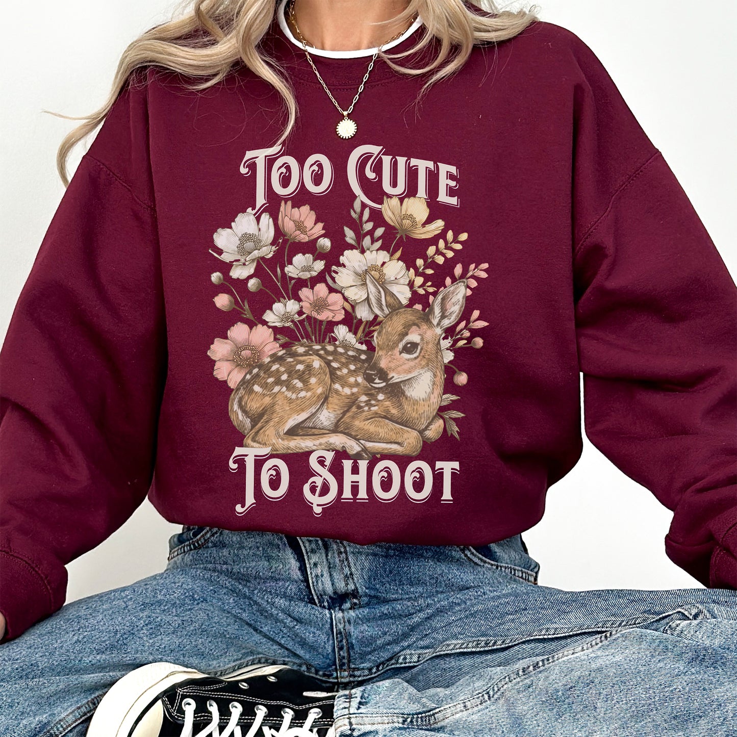 Maroon sweatshirt with a deer and floral design and text 'Too Cute To Shoot' worn by a woman in denim jeans.