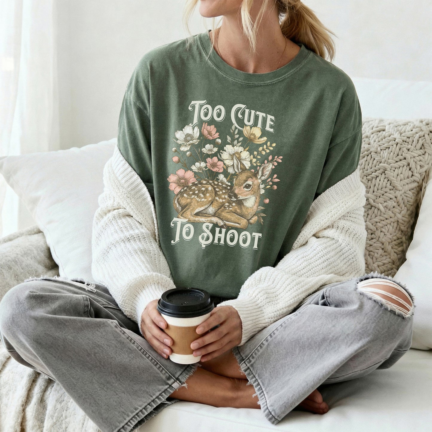 Person wearing a green tee shirt with a graphic design of a deer with the text too cute to shoot while sitting on a couch and holding a coffee cup.
