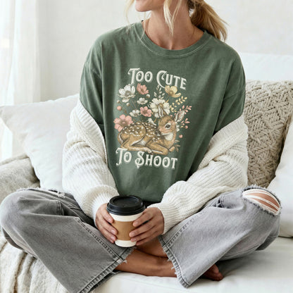 Person wearing a green tee shirt with a graphic design of a deer with the text too cute to shoot while sitting on a couch and holding a coffee cup.