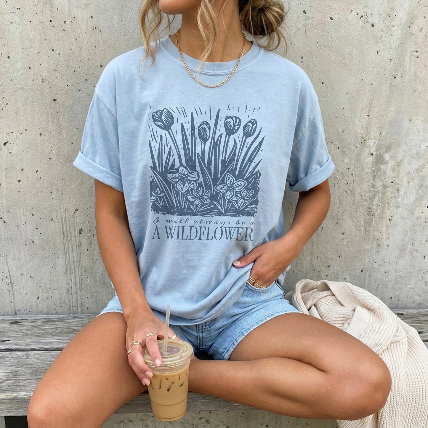Woman wearing a light blue t-shirt with floral design and text that reads I will always be a wildflower, sitting on a wooden bench.