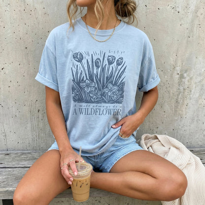 Woman wearing a light blue t-shirt with floral design and text that reads I will always be a wildflower, sitting on a wooden bench.